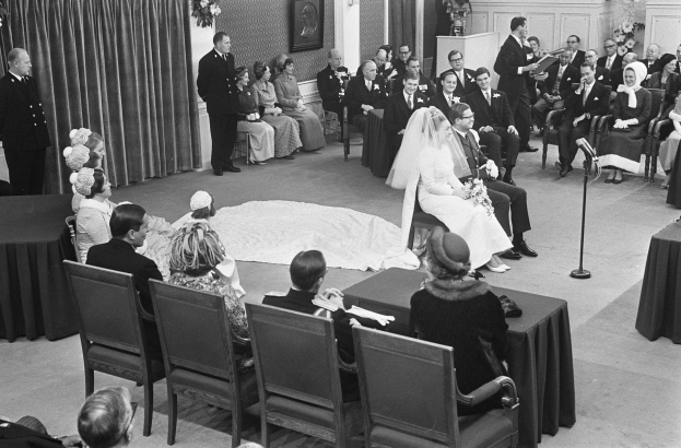 Schwarzes und weißes Foto einer Hochzeitszeremonie mit sitzenden und stehenden Menschen, ein Mann spricht in ein Mikrofon, eine Frau hält einen Blumenstrauß, ein Podium mit einem Mikrofon auf der rechten Seite und Blumenschmuck auf der linken Seite.