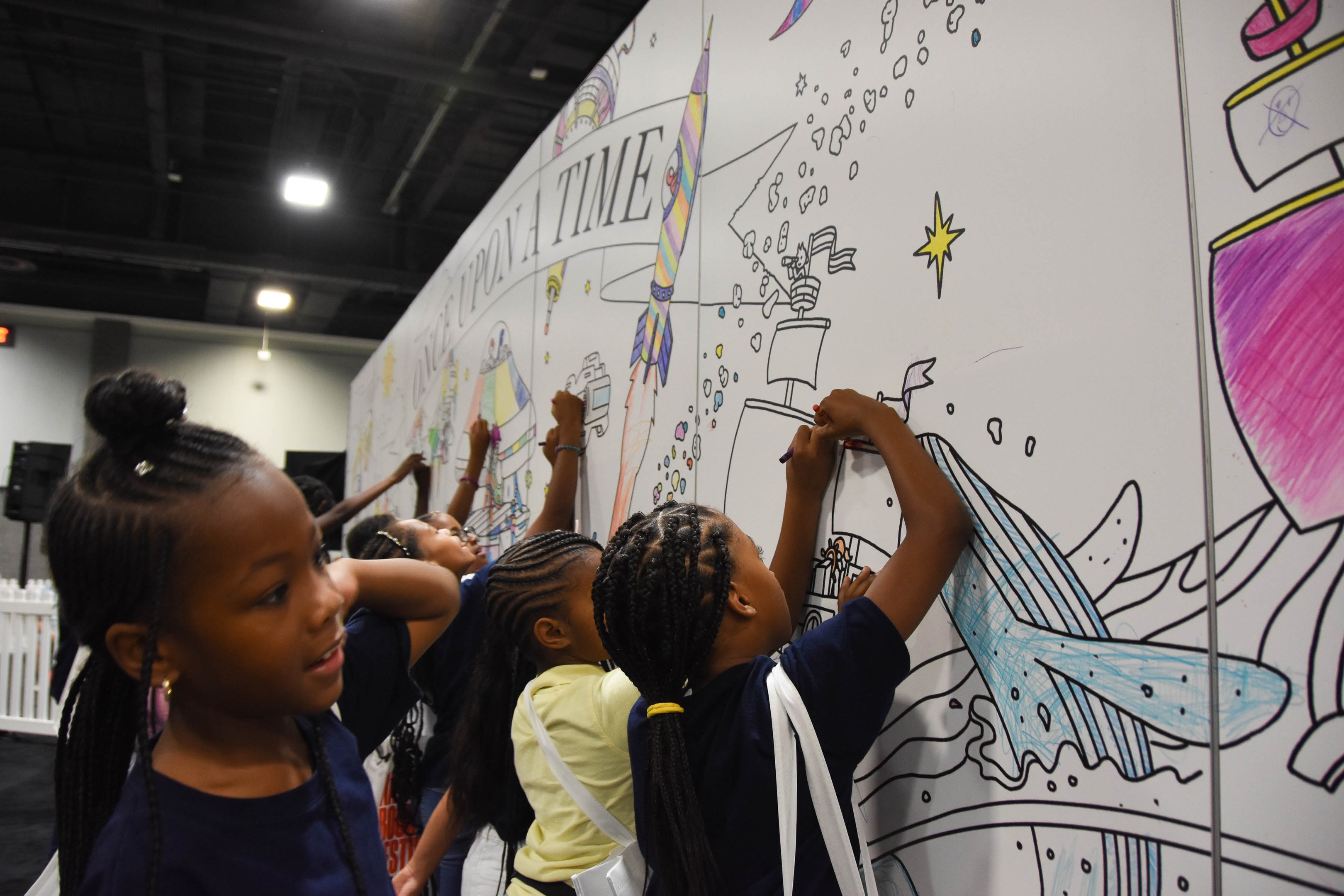 Kinder malen auf einer großeen Whiteboard mit Markern während einer Kunstausstellung, umgeben von Taschen, einem Zaun, Deckenlampen und Lautsprechern.