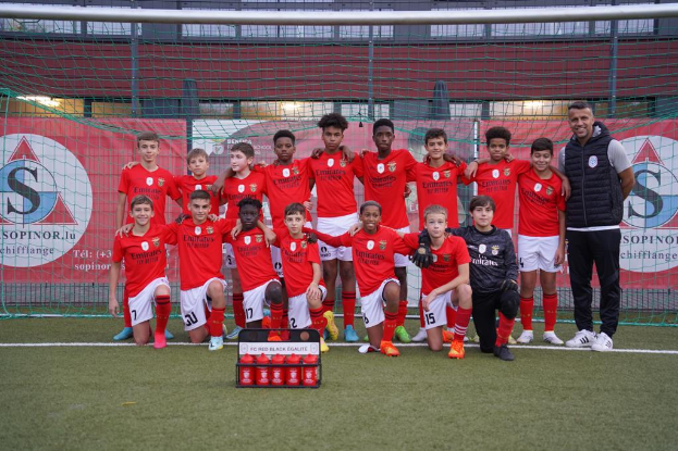 Eine Gruppe junger Männer in roten T-Shirts und schwarzen Shorts steht auf einem Fußballfeld vor einer Kiste roter Gegenstände, im Hintergrund Schilder mit der Aufschrift "FC Rot-Schwarz Eglaute - Champions League"
