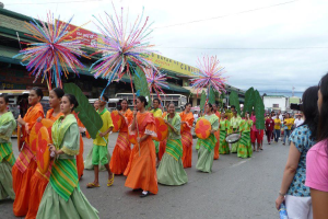 Eine Gruppe von Menschen in farbenfrohen Kostümen marschiert auf einer Straße während eines Karnevalsumzugs, mit einer Frau auf der rechten Seite, die eine Flasche hält, und Fahrzeugen, Gebäuden, Bäumen, Pfählen, Drähten und dem Himmel im Hintergrund.
