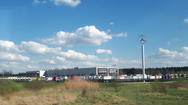 Ein Mercedes-Benz-Werk in Deutschland mit Fahrzeugen auf der Straße, umgeben von Gras, Pflanzen, Bäumen, Gebäuden und Polen, von denen einer ein Logo trägt, unter einem bewölkten Himmel.