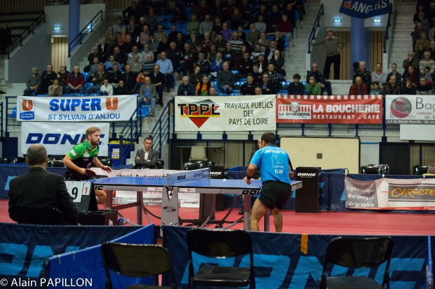 Indoor-Tischtennis-Match mit zwei Spielern, einigen stehenden Zuschauern, sitzendem Publikum, leeren Stühlen und Werbetafeln in einer Stadionatmosphäre.