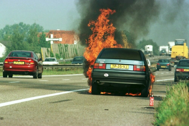 Ein Auto in Flammen auf der Straße, umgeben von anderen Fahrzeugen, mit Bäumen, Gebäuden und einem klaren blauen Himmel im Hintergrund und einem Feuerlöscher auf der rechten Seite.