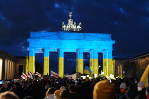 Eine Menschenmenge steht vor dem Brandenburger Tor in Berlin und hält Fahnen und Plakate, mit einer Banner auf der rechten Seite des Bildes.