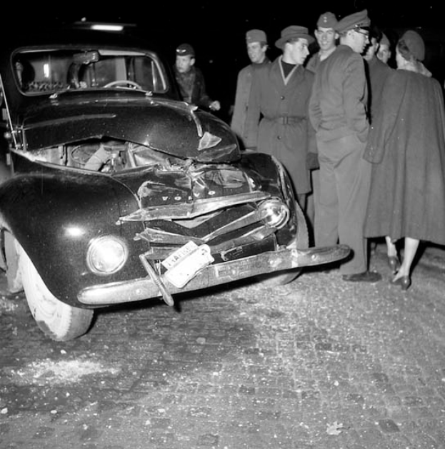 Ein Schwarz-Weiß-Bild eines verunglückten Autos am Straßenrand mit eingedellter Motorhaube und zersplitterten Fenstern, umgeben von einer Gruppe besorgter Menschen.