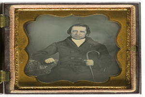 Ein Mann in einem Anzug sitzt in einem Stuhl und hält einen Stock in der Hand, mit einem ernsten Gesichtsausdruck und konzentriertem Blick in die Ferne, eingefangen in einem alten Daguerreotyp.
