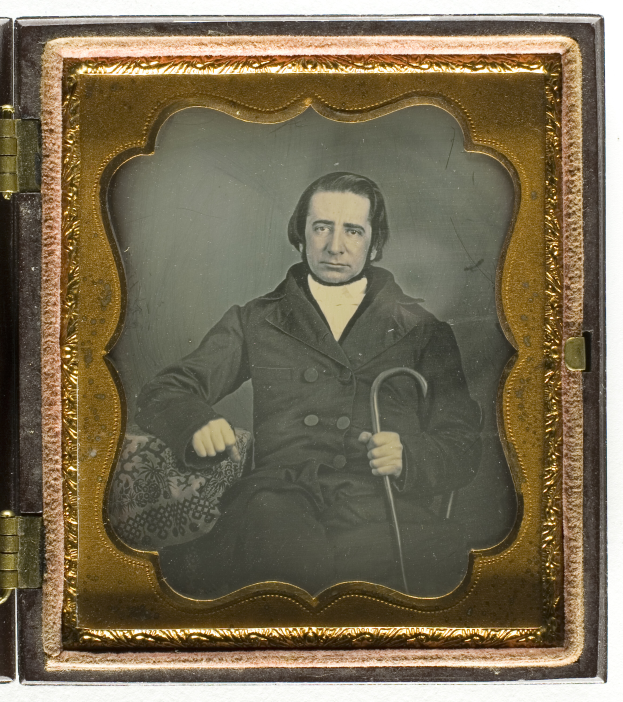 Ein Mann in einem Anzug sitzt in einem Stuhl und hält einen Stock in der Hand, mit einem ernsten Gesichtsausdruck und konzentriertem Blick in die Ferne, eingefangen in einem alten Daguerreotyp.