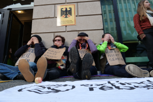 Eine Gruppe von Menschen sitzt vor einem Gebäude auf dem Boden, hält Schilder, mit einem Banner und einem Apfel auf der Straße, und eine Frau steht rechts daneben und sieht besorgt aus.
