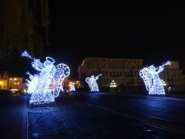 Eine Gruppe von Menschen, die nachts eine Straße entlanggehen, beleuchtet von Weihnachtslichtern, mit Skulpturen von Musikern im Vordergrund.