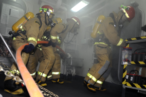 Feuerwehrleute in Schutzausrüstung und Helmen arbeiten an einem Feuerlöschwagen mit Sauerstoffflaschen, während ein Gestell mit Gegenständen und Hintergrundausrüstung zu sehen ist.
