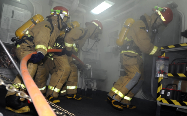 Feuerwehrleute in Schutzausrüstung und Helmen arbeiten an einem Feuerlöschwagen mit Sauerstoffflaschen, während ein Gestell mit Gegenständen und Hintergrundausrüstung zu sehen ist.