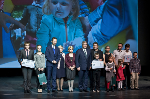 Eine Gruppe von Menschen auf einer Bühne mit Taschen und Urkunden, im Hintergrund ein Bildschirm mit einem Kind und anderen.