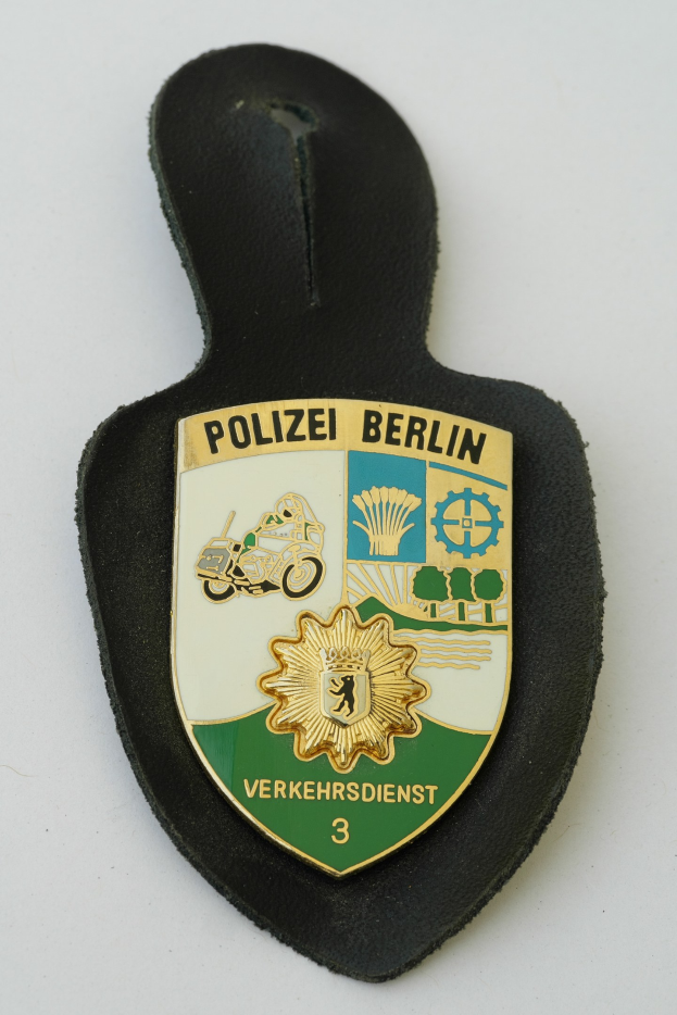Ein schildförmiges Polizeibadged mit "Polizei Berlin" in schwarzer, fetter Schrift auf einem blauen Hintergrund mit einem weißen Stern und Rahmen, das auf einer Oberfläche ruht.