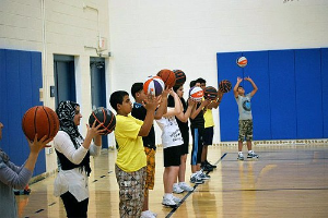 Gruppe junger Menschen mit Basketballs auf einem Court bei einem Basketballcamp, mit Türen und einer Wand im Hintergrund.