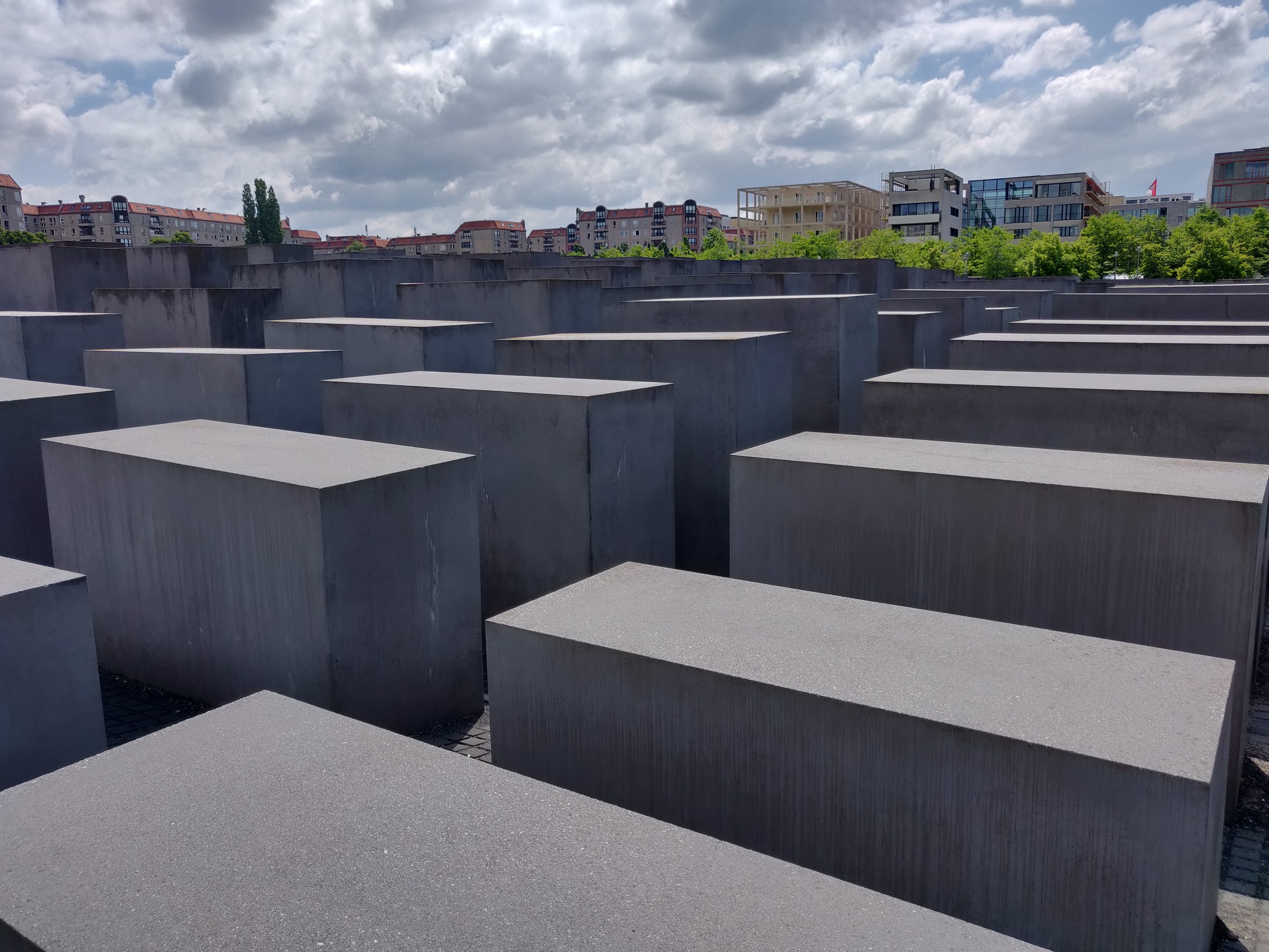 Betonschollen in einem Gittermuster am Holocaust-Mahnmal für die ermordeten Juden Europas in Berlin, mit Bäumen, Gebäuden und Wolken im Hintergrund.