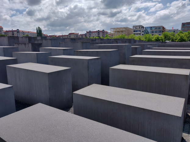 Betonschollen in einem Gittermuster am Holocaust-Mahnmal für die ermordeten Juden Europas in Berlin, mit Bäumen, Gebäuden und Wolken im Hintergrund.