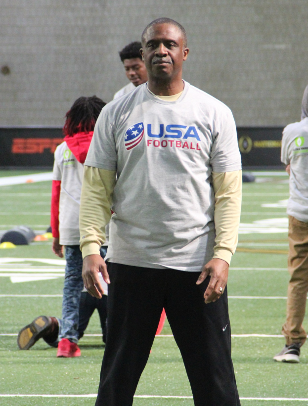 Ein Fußballspieler in Fußballschuhen steht auf einem Feld mit einer Gruppe von Menschen in der Nähe und einer Wand im Hintergrund.