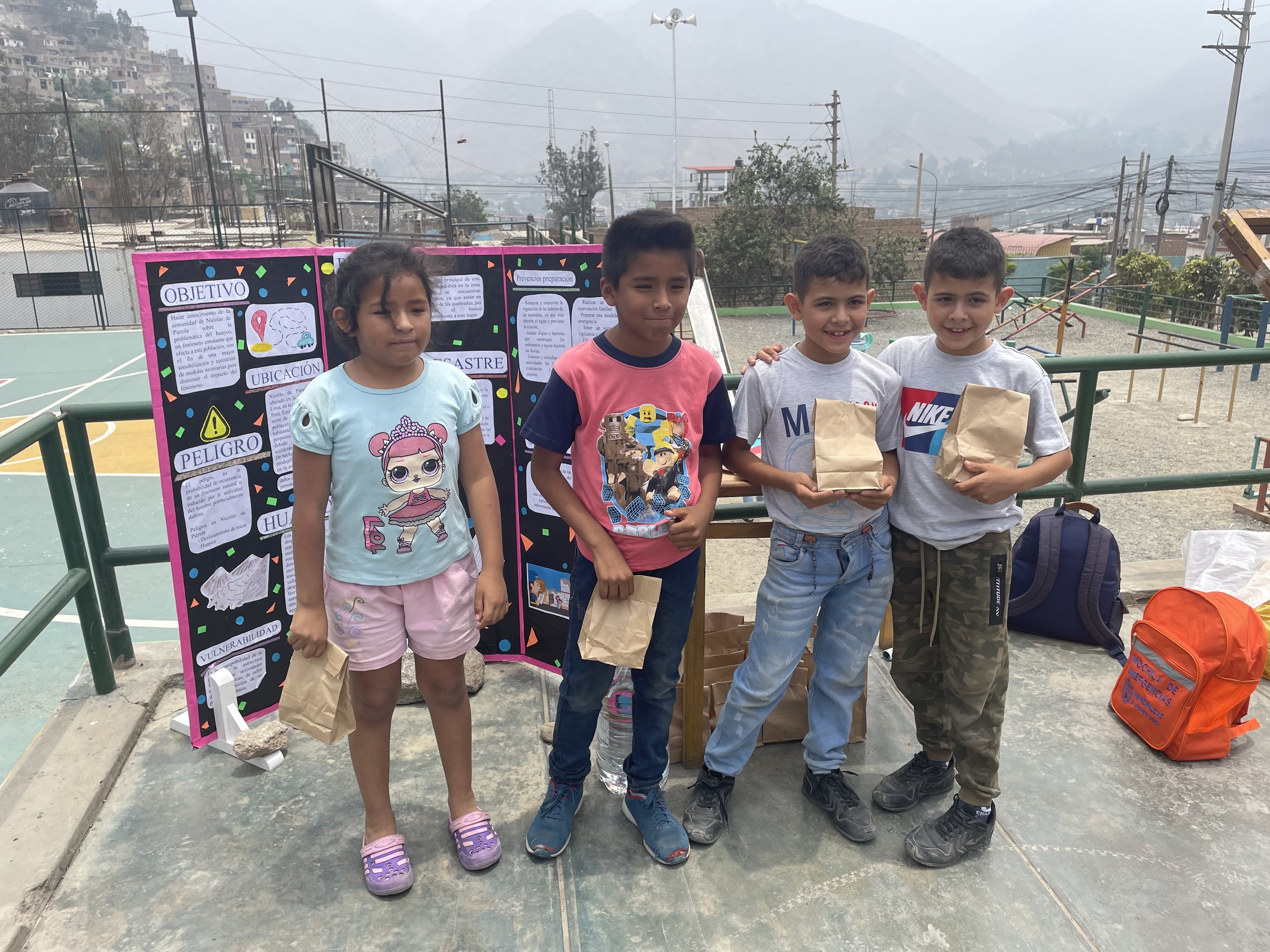 Eine Gruppe von Kindern steht auf einem Basketballfeld und hält Papiertüten in den Händen, mit Tüten und Gegenständen rechts und einer Tafel mit Text links, vor einem Hintergrund aus Zäunen, Pfosten, Laternen, Bäumen, Gebäuden, Hügeln und Himmel.