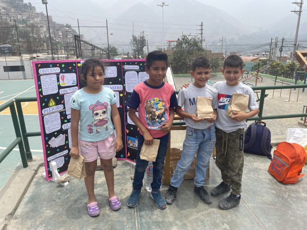 Eine Gruppe von Kindern steht auf einem Basketballfeld und hält Papiertüten in den Händen, mit Tüten und Gegenständen rechts und einer Tafel mit Text links, vor einem Hintergrund aus Zäunen, Pfosten, Laternen, Bäumen, Gebäuden, Hügeln und Himmel.