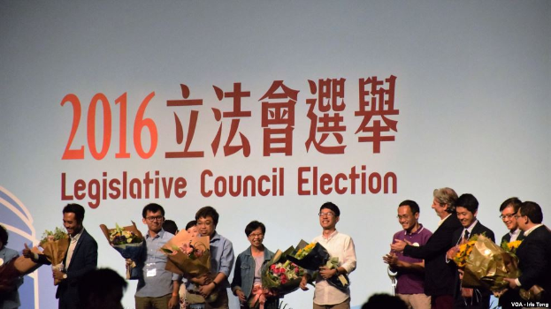 Eine Gruppe von Menschen auf einer Bühne mit Blumensträußen, darunter ein Publikum und ein Hintergrundbild mit Text und Logo zur Wahl des Legislativrats 2016.