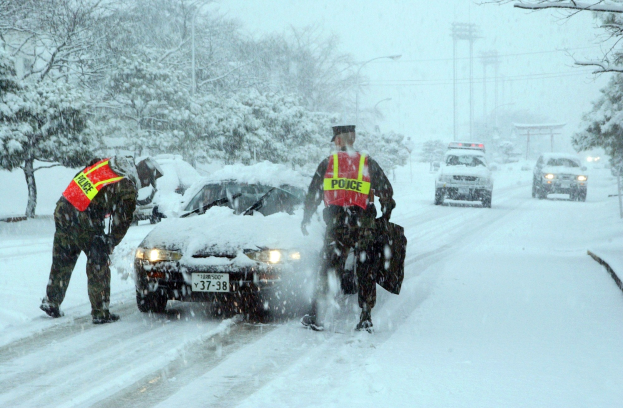 Zwei Polizeibeamte in Winterkleidung stehen neben einem stark schneebedeckten Auto auf einer verschneiten Straße, mit Bäumen, Strommasten und Drähten im Hintergrund unter einem klaren Himmel.