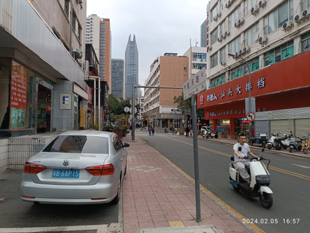 Ein Mann fährt mit einem Roller auf einer von Hochhäusern gesäumten Stadtstraße, mit Fußgängern auf den Gehwegen und anderen Fahrzeugen auf der Straße, unter einem klaren blauen Himmel mit Bäumen im Hintergrund.