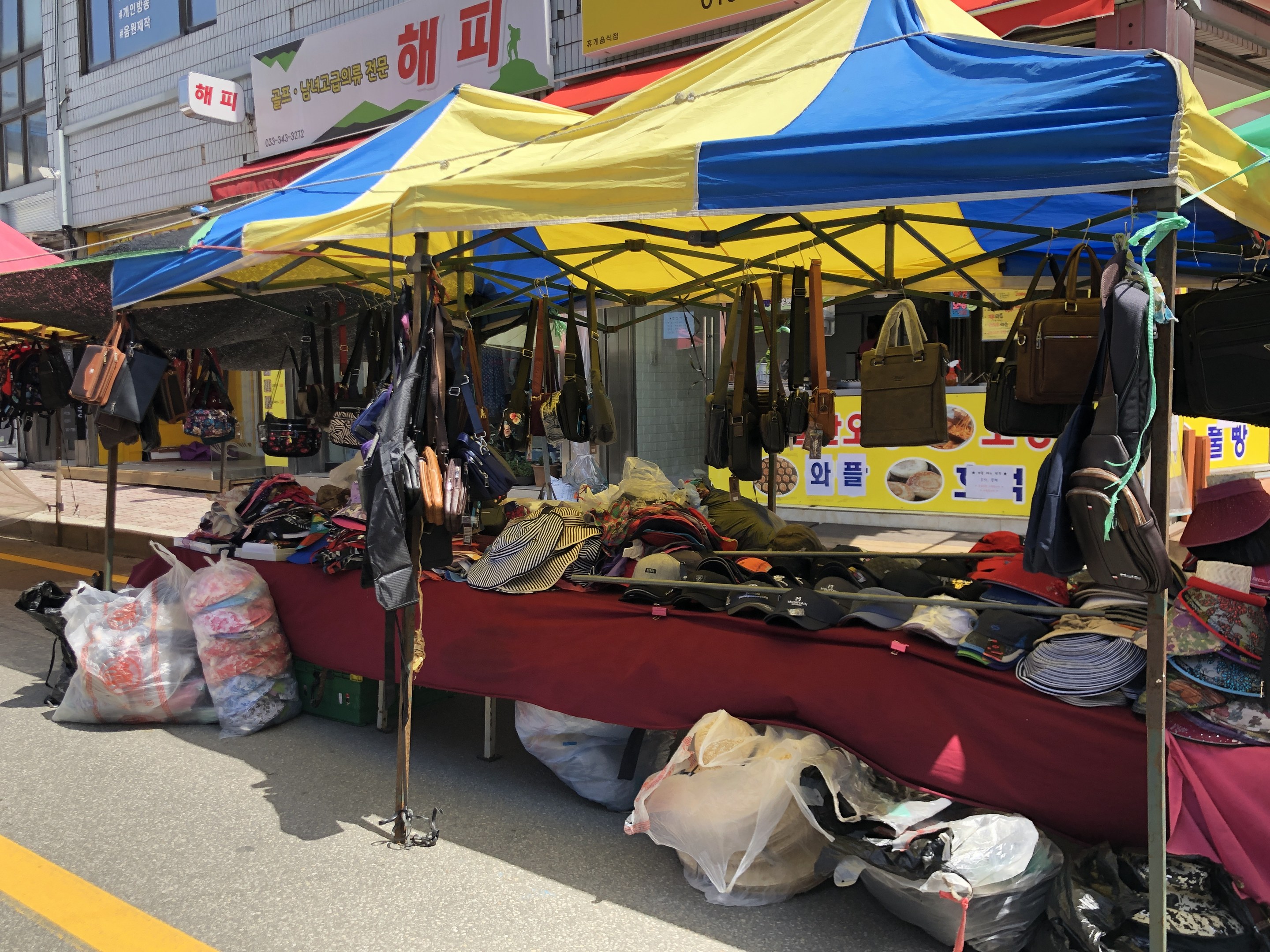 Ein Straßenmarkt mit Zelten, die Taschen, Hüte und andere Artikel präsentieren, vor Gebäuden mit Fenstern und Texttafeln.