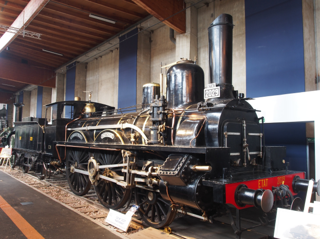 Alte Dampflokomotive in einem Museums Setting mit umgebenden Eisenbahnschienen, Informationsschildern und anderen Ausstellungen an einer Wand mit Deckenleuchten.