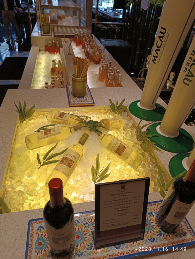 Ein Tisch mit Weinflaschen und Eiswürfeln, umgeben von Pflanzen und anderen Gegenständen, vor dem Hintergrund von Menschen, Tischen, Stühlen und Glastüren beim Macau-Bierfestival.