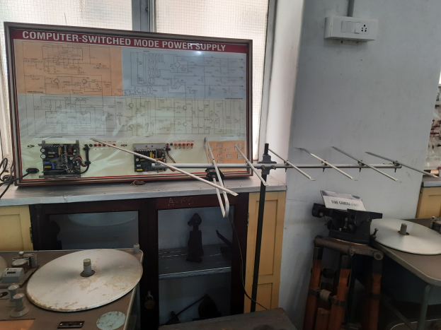 Ein unordentlicher Raum mit verschiedenen Elektronikgeräten, einschließlich einer beschrifteten Platine auf einem Tisch, Metallstangen, einer wandmontierten Schalttafel, einem Fenster und Gegenständen in Schränken, die einem Computer-ge-switchten Modus-Stromversorgungsetup ähneln.