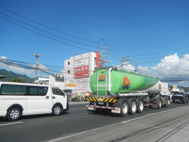 Ein großer Tanklastzug fährt auf einer Straße neben einem weißen Van, mit Strommästen, Gebäuden, Werbetafeln, Bäumen, Hügeln und einem bewölktem Himmel im Hintergrund.