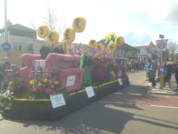 Ein Paradewagen, der mit Blumen und Pflanzen geschmückt ist und von Menschen begleitet wird, umgeben von Zuschauern, Schildern, Gebäuden, Bäumen und einem klaren blauen Himmel.