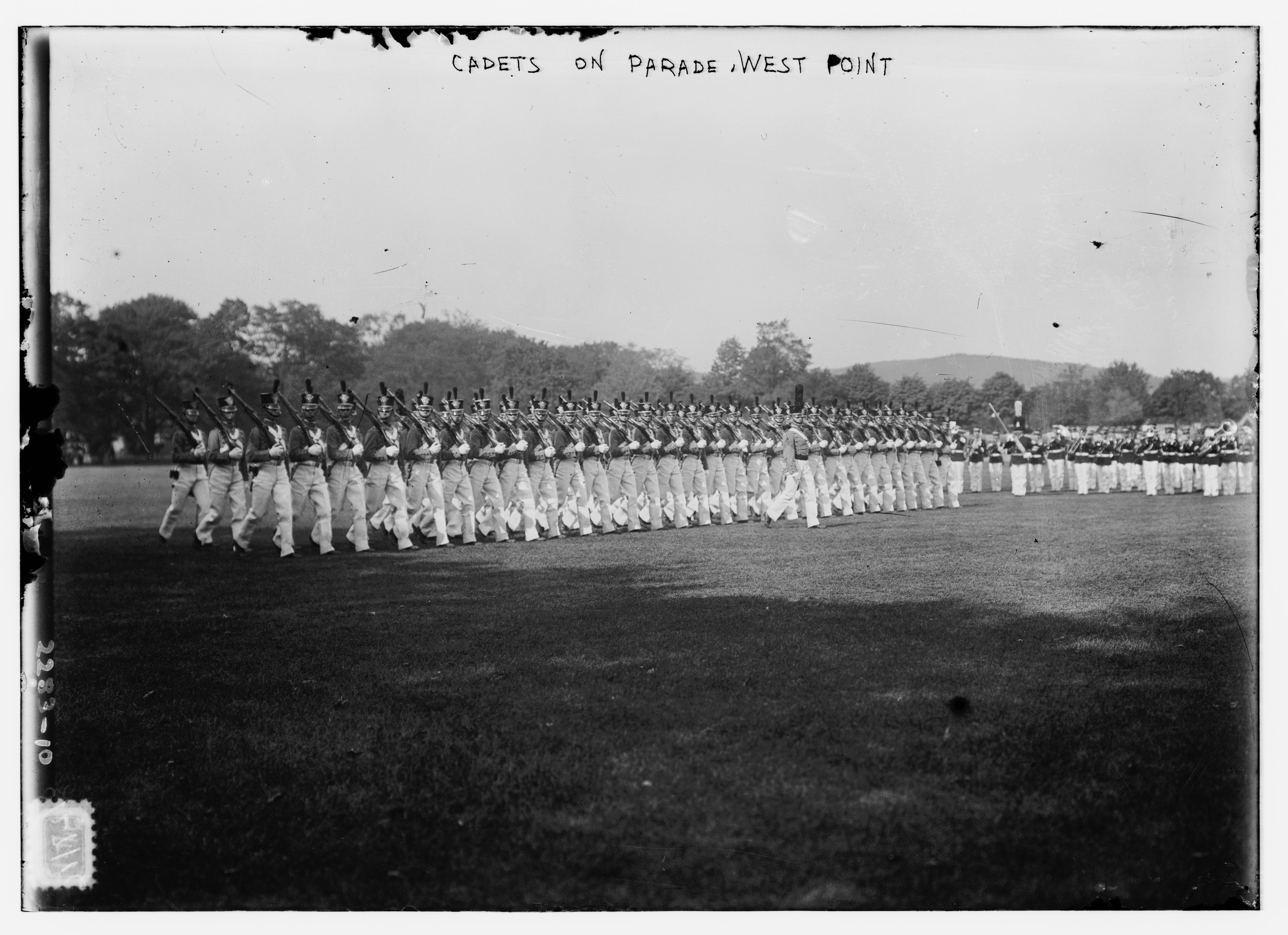 Gruppe von Menschen, einige mit Gewehren, auf dem Boden stehend mit Bäumen, Hügeln und einem bewölkten Himmel im Hintergrund, mit dem Text "cadets on parade west point" oben.