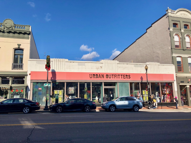 Eine belebte Stadtstraße mit parkenden Autos, Gebäuden mit Fenstern und einer "Urban Outfitters"-Reklame, Fußgängern auf dem Gehweg, Laternenmasten und einem bewölkten Himmel im Hintergrund.