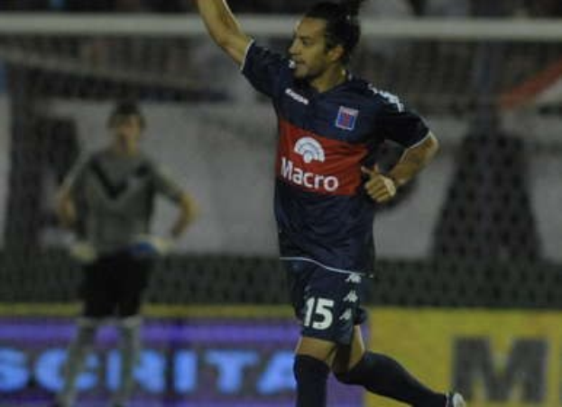 Ein Fußballspieler feiert auf dem Boden nach einem Tor, mit einer jubelnden Menge und einer Person im Hintergrund und einer Texttafel dahinter, alles vor einem leicht unscharfen Hintergrund.