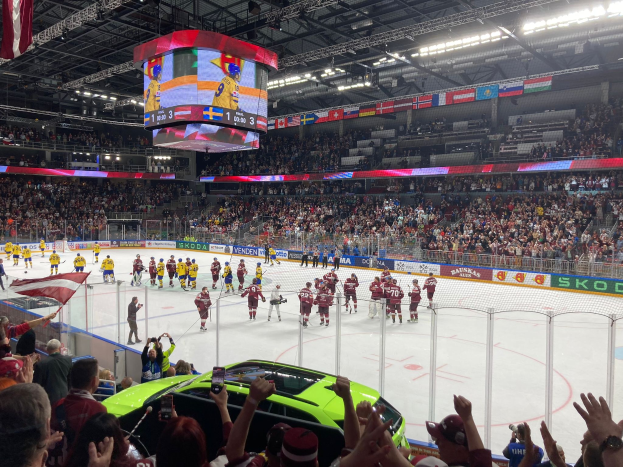 Ein Eishockeyspiel in einer großen Arena mit Zuschauern, eine Eisfläche umgeben von einem Zaun, Bannern, Deckenlampen und Bildschirmen sowie Flaggen an der Decke und in den Händen von Zuschauern, mit einem Auto im Vordergrund.