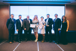 Gruppe von lachenden Menschen mit Urkunden vor einem Bildschirm, mit einem Tisch und einem wandmontierten Objekt im Hintergrund, Gewinner der International Business Awards 2018.