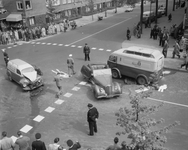 Schwarze und weiße Aufnahme von einem Autounfall an einer Straßenkreuzung mit mehreren Fahrzeugen, darunter ein Lieferwagen, und einer Gruppe von Menschen in der Nähe, mit Gebäuden, Bäumen, Pfählen und Texttafeln im Hintergrund.