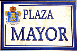 Rechtwinkliges weißes Schild mit schwarzer Schrift "Plaza Mayor" in fetter Schrift, blauem Rand und einem zentralen gekrönten Wappen-Logo, an einem Gebäude montiert.