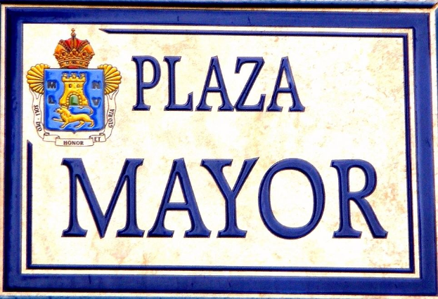 Rechtwinkliges weißes Schild mit schwarzer Schrift "Plaza Mayor" in fetter Schrift, blauem Rand und einem zentralen gekrönten Wappen-Logo, an einem Gebäude montiert.