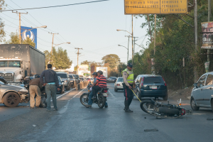 Eine Gruppe von Menschen steht um ein verunglücktes Motorrad auf der Straße herum, umgeben von mehreren Fahrzeugen, darunter ein Lastwagen, und einer Hintergrundkulisse aus Bäumen, Pfählen, Lampen und Schildern unter dem Himmel.
