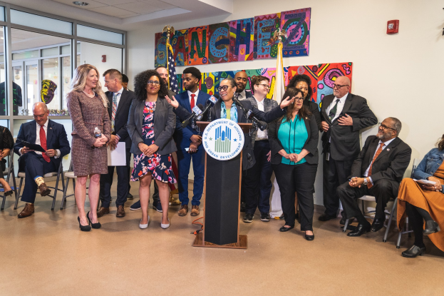 Gruppe von Menschen vor einem Podium mit Mikrofon und einer Tafel mit Text, mit sitzenden und stehenden Zuschauern, Flaggen, einem Feuerlöscher und Deckenleuchten im Hintergrund.