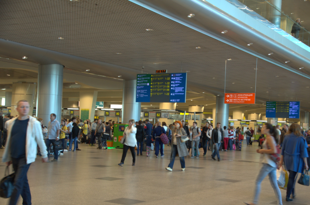 Menschen gehen durch einen Flughafen mit Überhangschildern, Säulen, Deckenleuchten und einem Glasgeländer auf der rechten Seite.