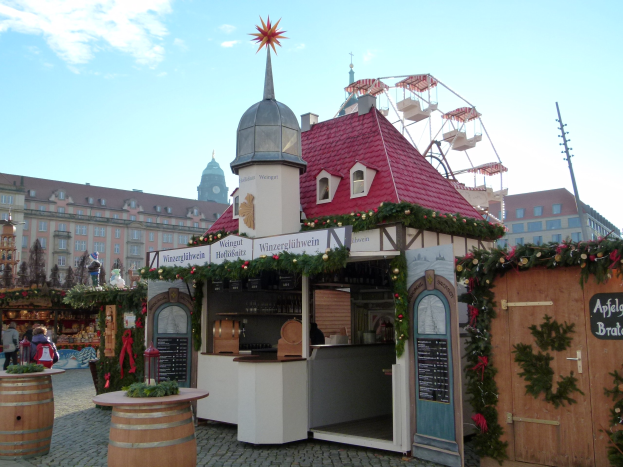 Ein kleines Gebäude mit einem roten Dach und einem Riesenrad davor, umgeben von Menschen und Festdekorationen bei einem Outdoor-Event mit Gebäuden, Bäumen und einem bewölkten Himmel im Hintergrund.