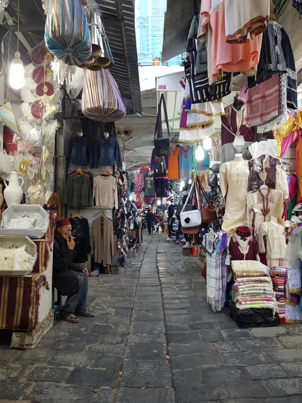 Ein belebter Straßenmarkt in der Altstadt von Jerusalem mit Ständen, die Kleidung, Taschen, Lampen und andere Artikel verkaufen, während Menschen im Vordergrund durch die Stände schlendern und interagieren, mit einem Gebäude im Hintergrund.