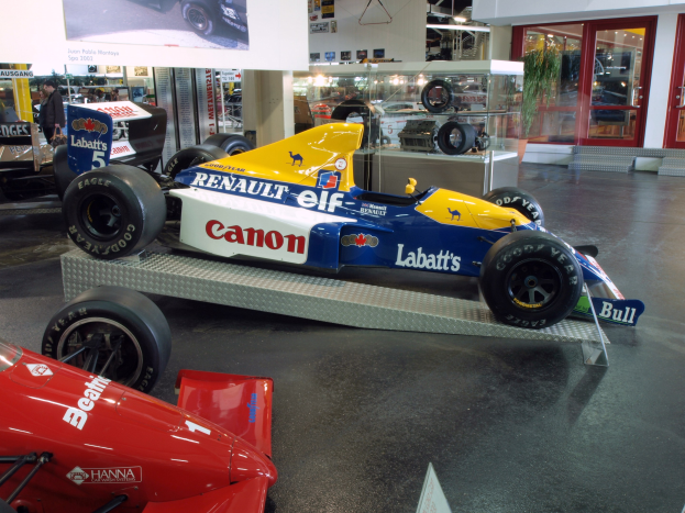 Eine Museumsausstellung zeigt verschiedene Rennwagen, darunter ein Renault-Formel-1-Auto, mit Besuchern, die beobachten, und einer Texttafel an der Wand unter beleuchteten Decken und Glastüren.