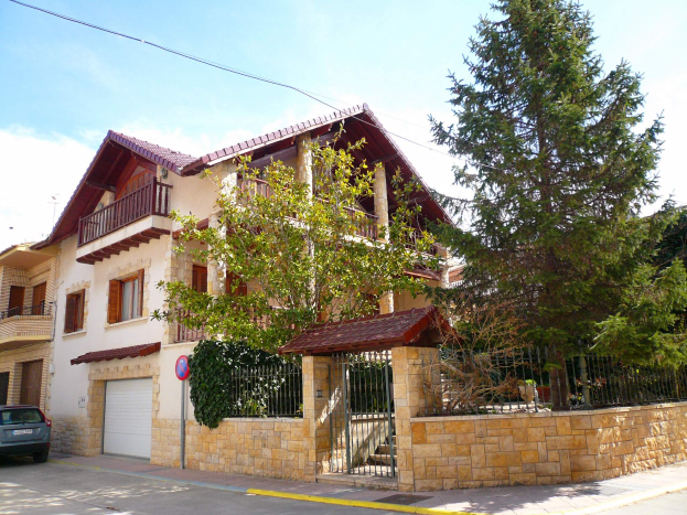 Eine Villa mit einem vor ihr geparkten Auto, das Fenster, Geländer, Pfeiler, ein Dach und eine Mauer hat, umgeben von einem Tor, einem Schild, einem Straßenschild, Bäumen und einer Wand, unter einem bewölkten Himmel.