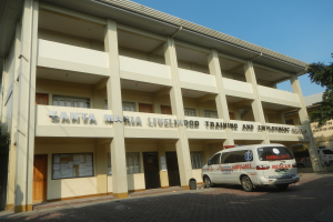 Santa Maria Lebensmittelpunkt- und Arbeitsst├Ątte-Geb├Ąude mit Fenstern, Türen und Text überwachsen von B├Ąumen und Fahrzeugen auf der Stra├če unter einem klaren blauen Himmel.
