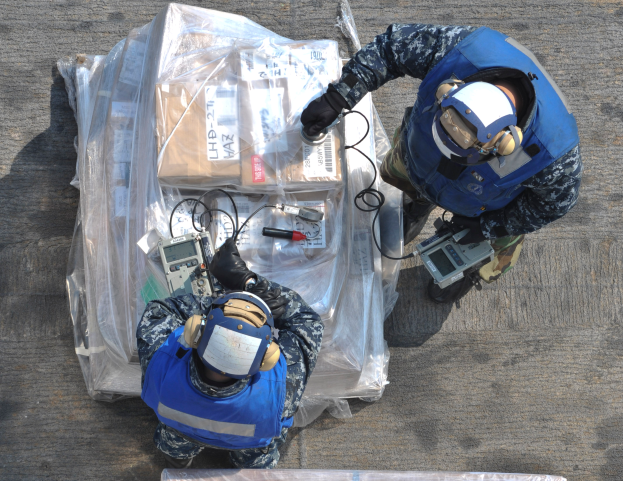 Zwei Männer in blauen Uniformen und Helmen arbeiten an einem großen Drohnenmodell, halten verschiedene Gegenstände und haben eine Plastikabdeckung vor sich auf dem Boden.