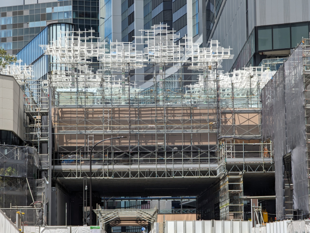Großes Gebäude mit Gerüst in einer städtischen Umgebung, umgeben von Bäumen auf der linken Seite und einer Straße unten.